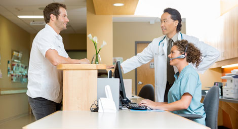 Man stood at a reception desk with a receptionist seated opposite with a clinician stood to the side of her